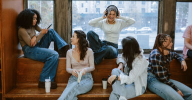 A mixed-raced group of young women sit on a bench and ledge interacting.