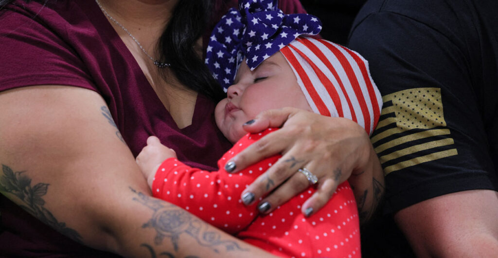 A dark haired mother with tattooed arms holds an infant dressed in red-and-white with its head wrapped in an American flag cap.