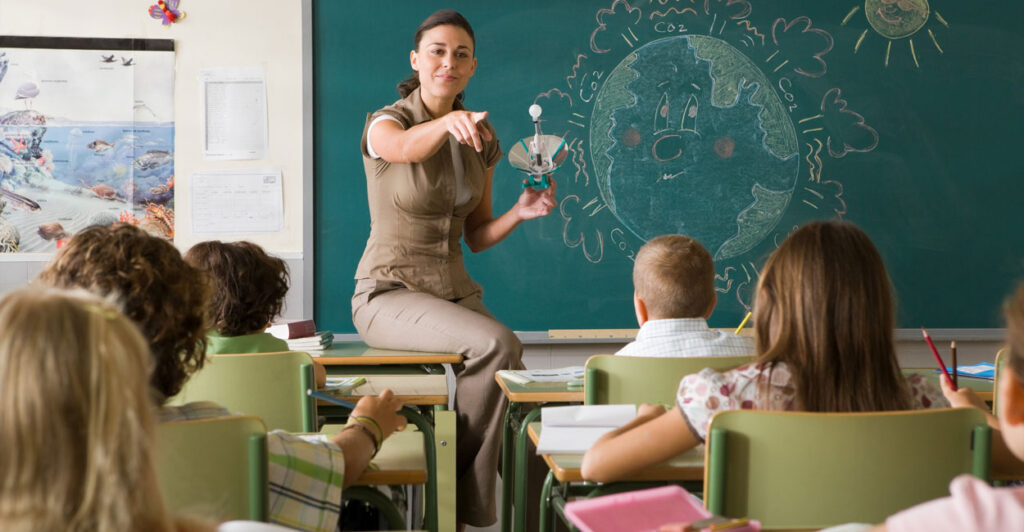 A female teacher sits on a desk in front of a blackboard pointing toward her students.