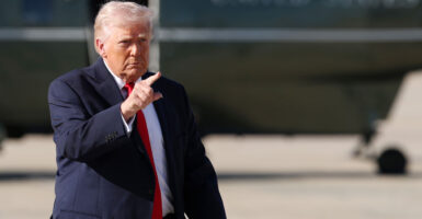 Donald Trump walking across the tarmac with Marine One in background pointing.