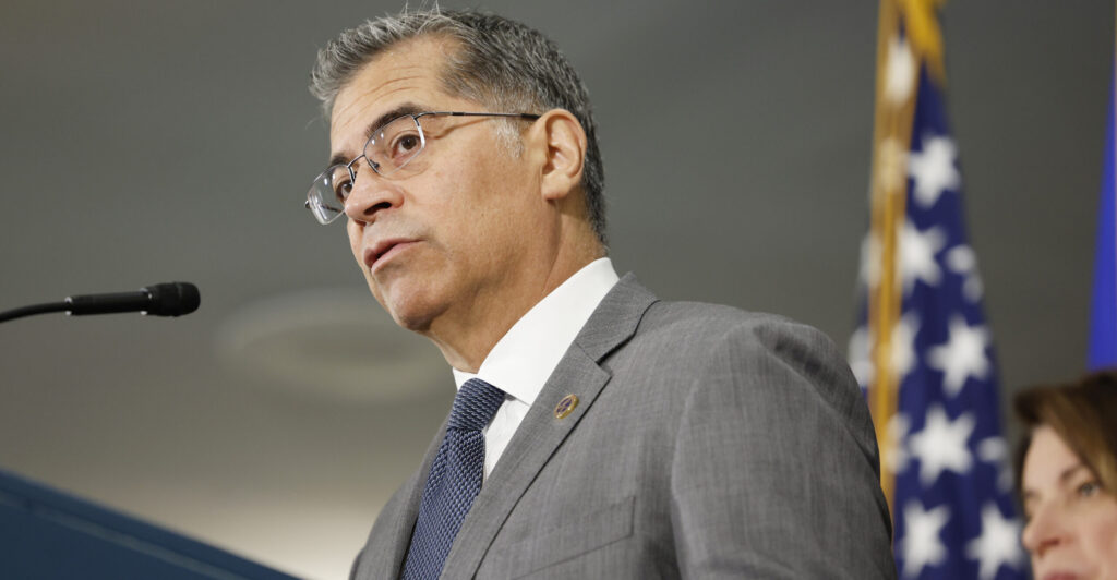 Health and Human Services Secretary Xavier Becerra stands at a podium with an American flag in the background.