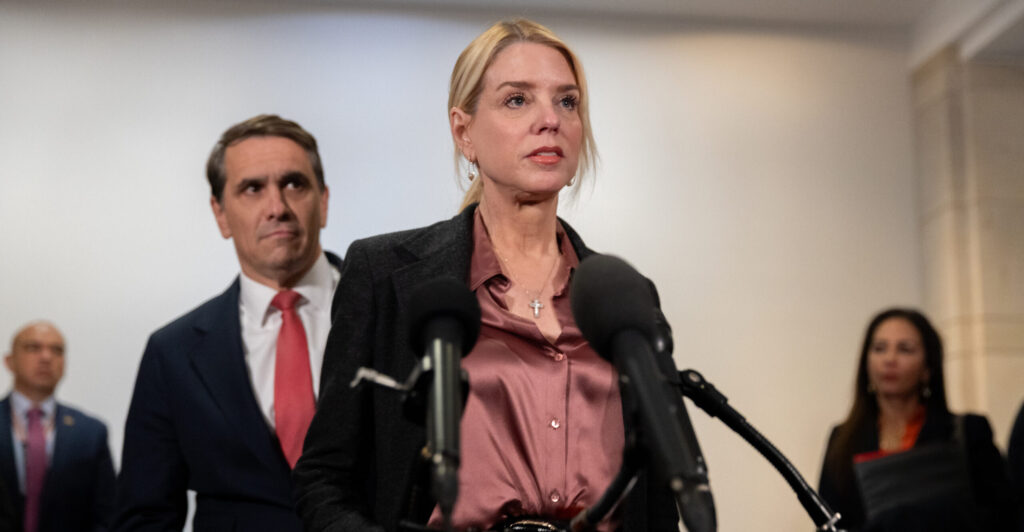 Pam Bondi in a purple blouse and black suit talks to reporters with Todd Blanche behind her.